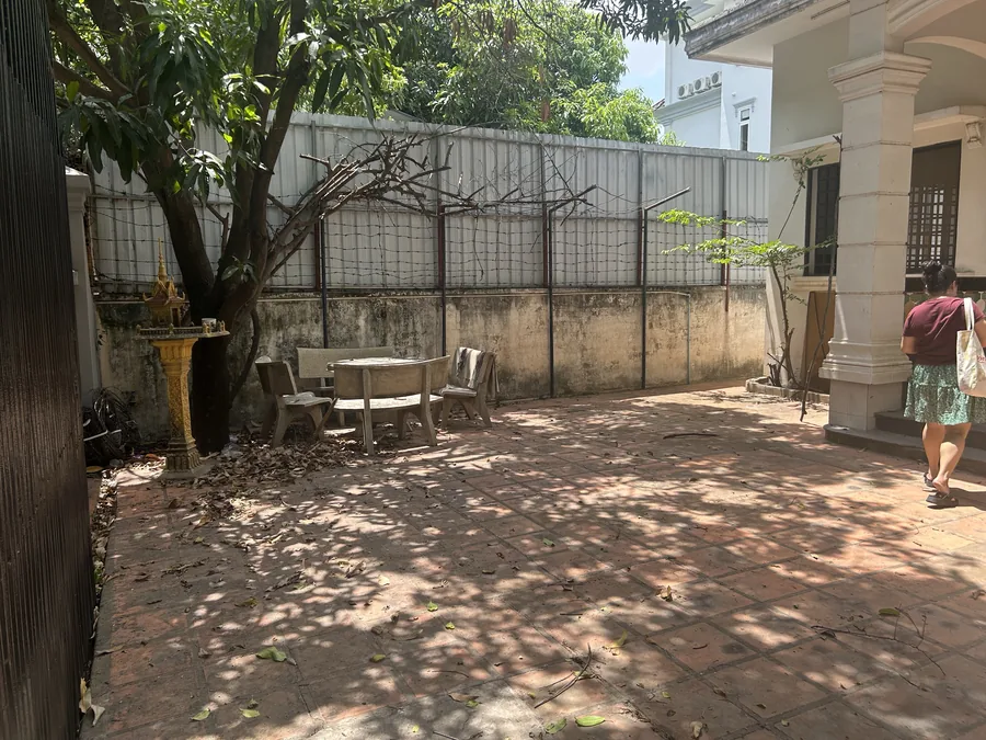 The courtyard before renovation: bare brick paving, a weathered wall with barbed wire, the mango tree, stone table and benches, and the gold spirit house