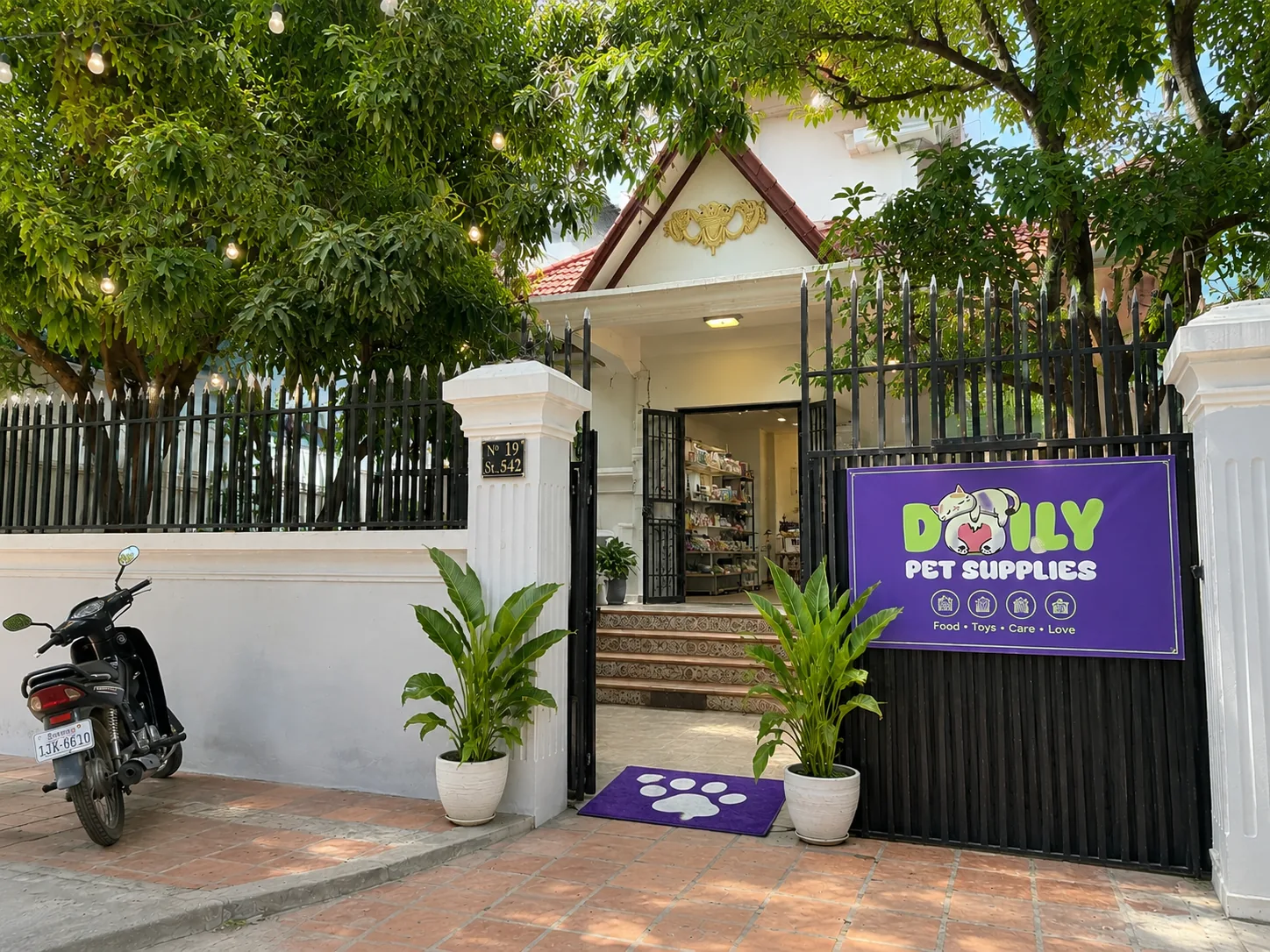 The gate of Daily Pet Supplies: a white villa with a red-tiled gable, black iron fence, big shade trees and a purple shop sign